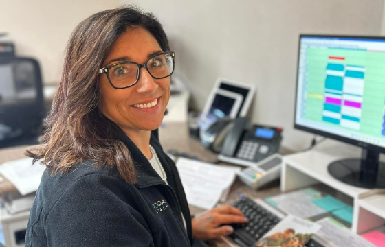 Smiling woman with glasses at a desk, typing on a keyboard. A computer screen with a colorful schedule is visible. Office setting, positive mood.